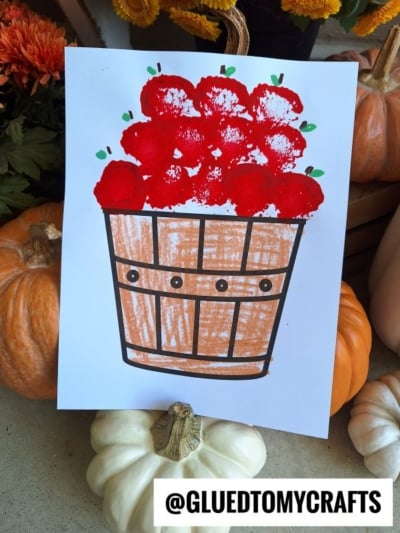 A handmade drawing of a wooden apple basket filled with red pom pom apples made from sponge or finger painting, surrounded by pumpkins and autumn decor. The handle @GLUEDTOMYCRAFTS is at the bottom.