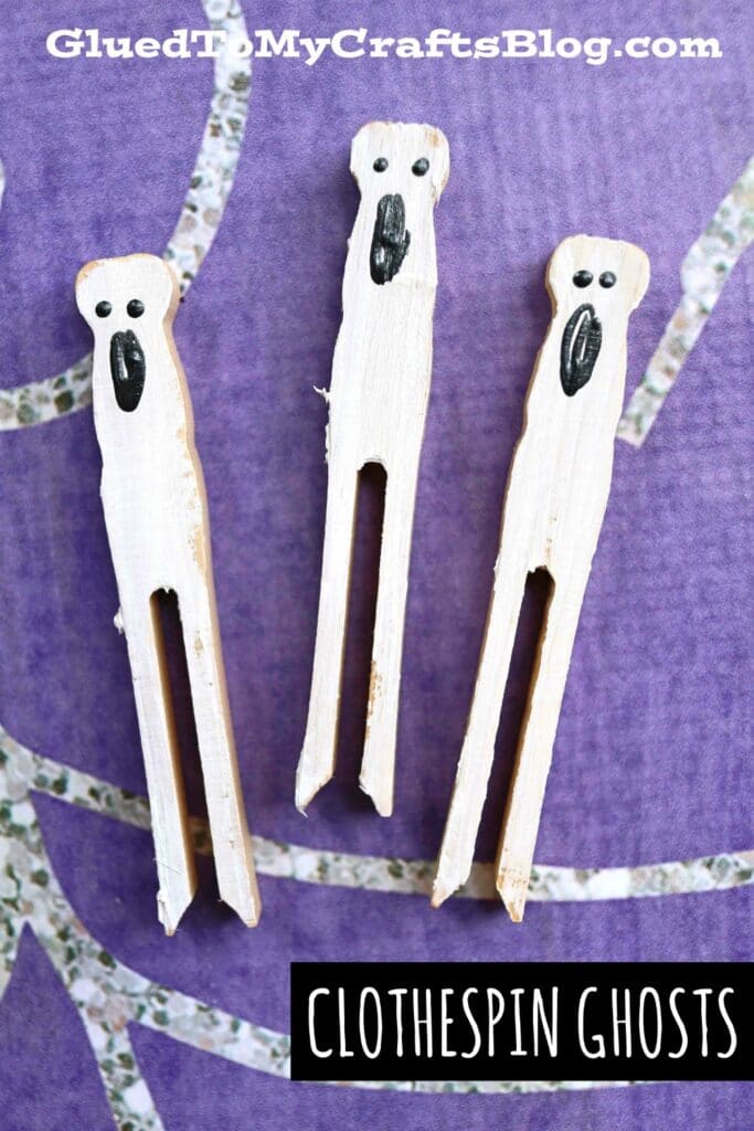 Three wooden clothespins painted white with ghost faces drawn in black are arranged on a purple background. Text at the bottom reads “CLOTHESPIN GHOSTS,” perfect for creating fun halloween clothespin puppets.