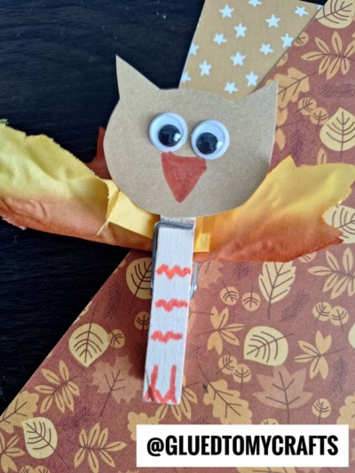 A handmade owl clothespins craft made with a clothespin, googly eyes, a paper beak, yellow and orange feathers, and patterned autumn-themed paper. The text @GLUEDTOMYCRAFTS appears at the bottom.