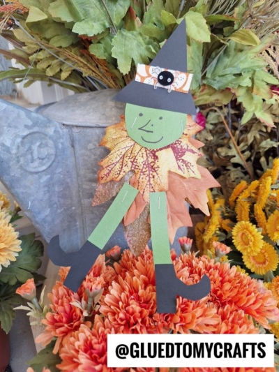 A cute leaf witch craft made from paper, green craft sticks, and autumn leaves sits among orange flowers and fall foliage. The witch has a black hat with a spider, a smiling face, and black boots. @GLUEDTOMYCRAFTS is at the bottom.