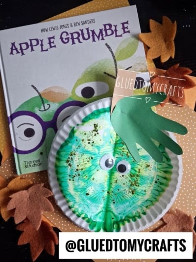 A children’s book titled Apple Grumble sits by a green paper plate apple craft with googly eyes and a green handprint leaf, surrounded by felt autumn leaves, paint splat apples, and a polka-dotted background.