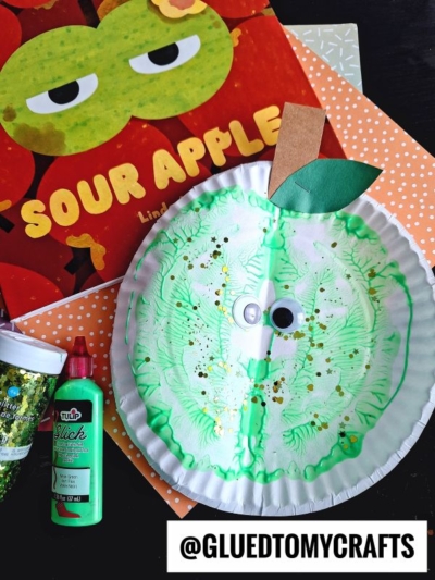 A green apple craft made from a paper plate with googly eyes, green paint, glitter, and paint splat apples is shown next to a Sour Apple book, green glitter, red paint, and colored paper. The handle @GLUEDTOMYCRAFTS appears at the bottom.