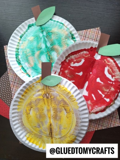 Three paper plate paint splat apples in green, red, and yellow with painted designs, brown paper stems, and green paper leaves are displayed on patterned craft paper. @GLUEDTOMYCRAFTS is written at the bottom.