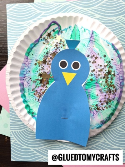 A paper plate peacock decorated with swirled marker colors and gold star confetti, topped with a blue paper peacock featuring big round eyes and a yellow beak. @GLUEDTOMYCRAFTS is written at the bottom.
