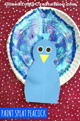 A marbled peacock craft made from a paper plate painted blue and purple to mimic feathers, topped with a blue peacock head and googly eyes, set on a red polka dot background. Text: “PAINT SPLAT PEACOCK.”.