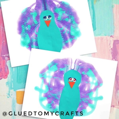 Two teal bird crafts with big eyes and purple-green textured wings are displayed on white paper, set against a colorful pastel painted background. Reminiscent of a marbled peacock craft, these charming creations bring whimsical flair to any art display. Text @GLUEDTOMYCRAFTS is at the bottom left.
