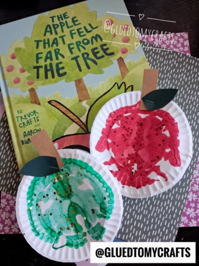 A children’s book titled The Apple That Fell Far From The Tree lies on a table with two vibrant paint splat apples crafted from paper plates, one red and one green, decorated with shimmering glitter and paper leaves. @GLUEDTOMYCRAFTS is labeled.