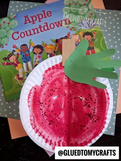 A paper plate painted red with black dots, reminiscent of paint splat apples, and topped with a green paper handprint leaf sits on books including Apple Countdown. A clothespin acts as the apple stem. @GLUEDTOMYCRAFTS watermark is present.