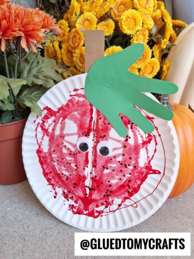 A paper plate decorated to look like a paint splat apple with red paint, googly eyes, a hand-shaped green leaf, and a brown stem, placed before artificial yellow flowers and a pumpkin. @GLUEDTOMYCRAFTS is written at the bottom.