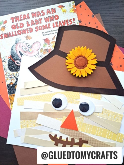 A children’s book, There Was an Old Lady Who Swallowed Some Leaves, lies next to orange and brown craft paper and a colorful paper scarecrow craft with a sunflower hat and googly eyes.