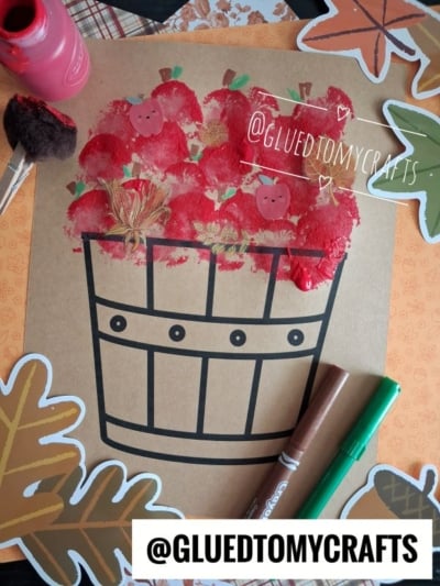 A paper with a printed apple basket design, red paint dabbed as pom pom stamped apples—some with drawn faces—surrounded by fall leaves, markers, paint, and a sponge. Text reads @GLUEDTOMYCRAFTS.
