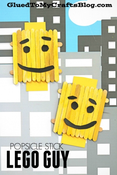 Two yellow popsicle stick Lego crafts shaped like smiling faces with drawn-on brows are displayed against a cityscape background. The text reads Popsicle Stick Lego Guy and GluedToMyCraftsBlog.com.