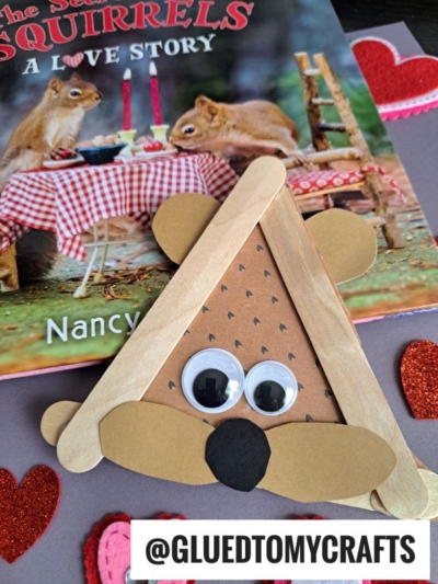 A squirrel-themed children’s book features a popsicle stick squirrel craft with felt heart decorations, googly eyes, and paper ears, nose, and cheeks. @GLUEDTOMYCRAFTS is labeled at the bottom.