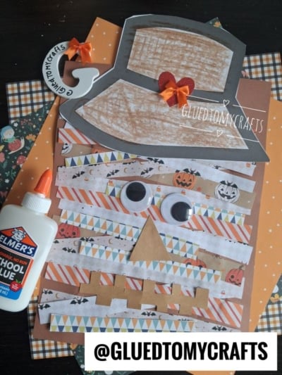 A Halloween-themed paper scarecrow craft made with patterned paper, googly eyes, a paper hat with a bow, and a glue bottle on the side. Background features orange, polka dot, and pumpkin papers. @GLUETOMYCRAFTS is labeled.