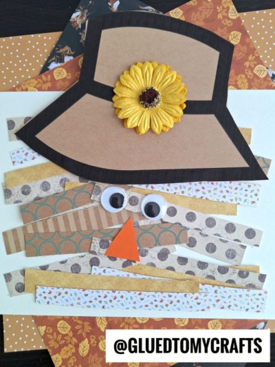 A paper scarecrow face made from layered patterned paper strips, a brown paper hat with a yellow flower, googly eyes, and an orange triangle nose. Autumn-colored papers are in the background. @GluedToMyCrafts is at the bottom.