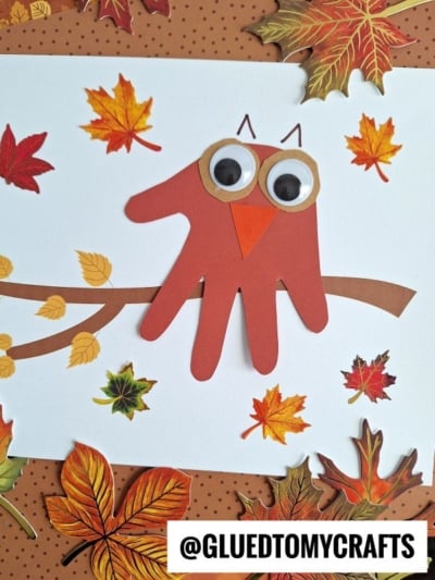 A Handprint Autumn Owl made from red paper sits on a branch, surrounded by colorful autumn leaves. The owl features large googly eyes and an orange beak, bringing this charming craft to life. A @GLUEDTOMYCRAFTS label is at the bottom.