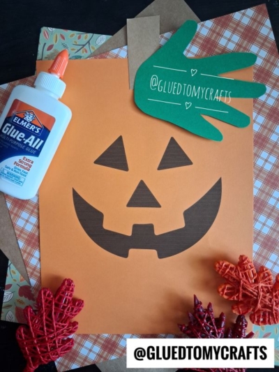Orange handprint jack-o’-lantern craft made from paper, surrounded by red leaves, plaid paper, a glue bottle, and a green cutout hand labeled “@GLUEDTOMYCRAFTS.” The username is also on a white label at the bottom.