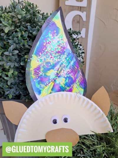A colorful gnome craft featuring a paper plate face, brown paper ears, googly eyes, and a tall hat decorated with glitter and vibrant paint splat troll hair. The handle @GLUEDTOMYCRAFTS appears in a green box.