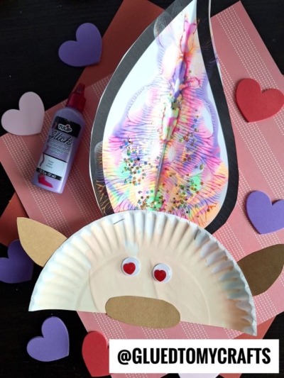 A paper plate craft resembling a troll face with googly eyes is displayed on pink and red paper adorned with heart shapes, glitter, and glue. The troll features a large, vibrant paint splat troll hairpiece for an extra burst of color.