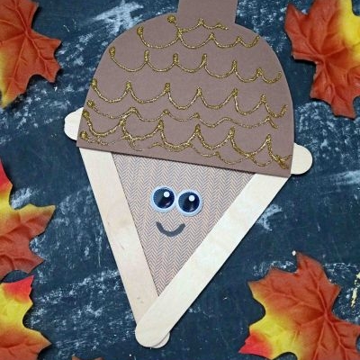A craft stick acorn made from brown paper, googly eyes, and craft sticks, decorated with gold glitter and surrounded by artificial autumn leaves on a dark background.
