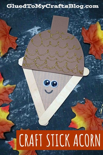 A craft stick acorn made from brown paper, googly eyes, and craft sticks, decorated with gold glitter and surrounded by artificial autumn leaves on a dark background.