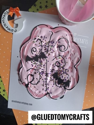 A butterfly-shaped paper features pink paint, purple and black glitter, and black bat stickers—perfect for sparking creativity. A decorated cup and orange polka dot paper appear in the background. For a fun twist, try adding puffy paint brains to your own design! Text reads @GLUETOMYCRAFTS and GLUETOMYCRAFTS.