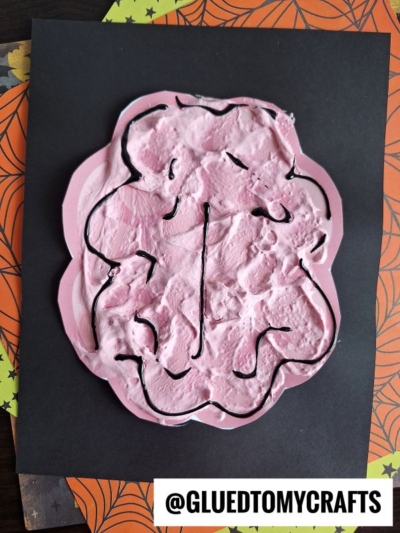 A pink, textured brain shape made with puffy paint brains and black squiggly lines is glued to black paper, resting on orange and green Halloween-themed paper. @GLUEDTOMYCRAFTS is printed at the bottom.