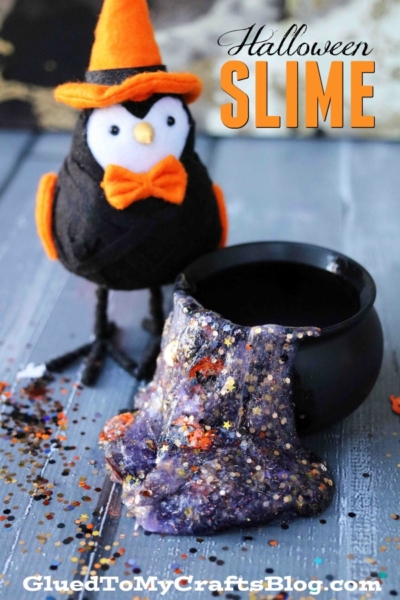 A small black cauldron overflows with sparkly purple magical liquid next to a black owl plush dressed in an orange witch hat and bow tie. The text “Halloween Slime” is at the top. Craft decorations are scattered around.