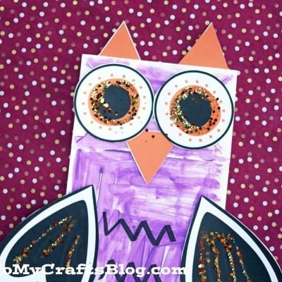 A colorful paper bag Halloween owl puppet made from craft materials, featuring big eyes, glittery wings, and a purple body, displayed on a maroon background with white polka dots.