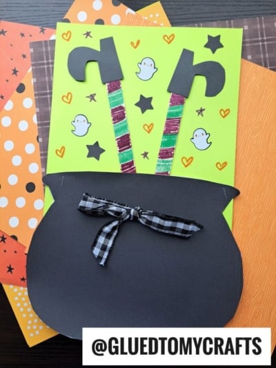 A Halloween craft with a black paper cauldron, striped popsicle stick witch legs sticking out, and a plaid bow. The background is green with doodles of stars, hearts, and ghosts, plus patterned paper sheets below.