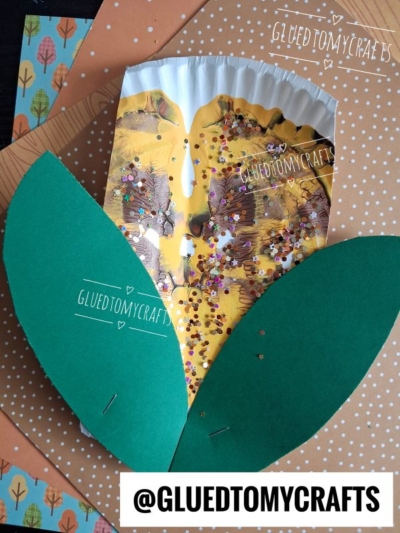 A corn craft inspired by paint splat indian corn, made from a painted, glittered paper plate with two green paper leaves, placed on patterned paper. The handle @GLUEDTOMYCRAFTS appears at the bottom.