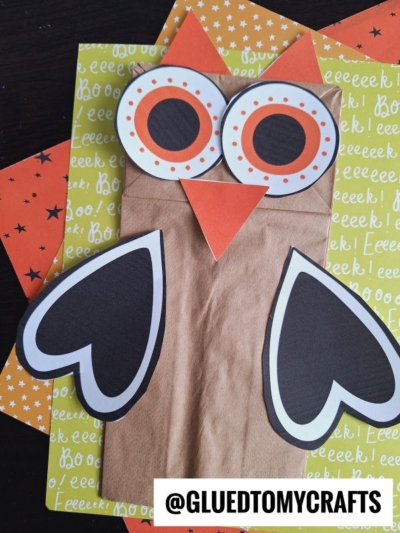 A paper bag halloween owl with big eyes, an orange beak, and heart-shaped wings sits on colorful patterned paper. This adorable paper bag owl craft makes the perfect halloween owl puppet. @GLUEDTOMYCRAFTS is written at the bottom.