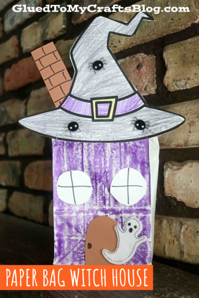 A paper bag witch house crafted with a purple door, white windows, a ghost, and a large gray witch hat with spider decorations, set against a brick wall—a spooky twist on the classic lunch sack haunted house craft.