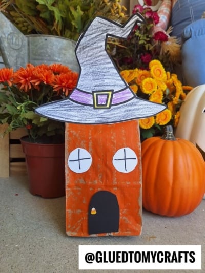 A Halloween craft featuring a rectangular orange block transformed into a paper bag witch house with drawn eyes and a door, topped with a large, hand-colored witch hat. Fall flowers, a pumpkin, and a watering can are in the background. Text: @GLUEDTOMYCRAFTS.