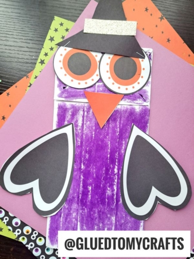 A purple popsicle stick craft shaped like an owl witch, with big orange and black eyes, black wings, a beak, and a hat—perfect inspiration for your own halloween owl puppet or paper bag halloween owl project! Displayed on colorful paper backgrounds. @GLUEDTOMYCRAFTS is at the bottom.
