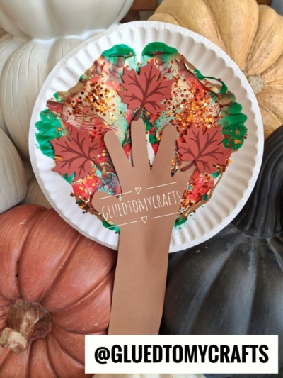 A paint splat tree craft made from a paper plate, featuring a brown paper hand cutout as the trunk and branches, adorned with glitter and paper leaves, and surrounded by decorative pumpkins.