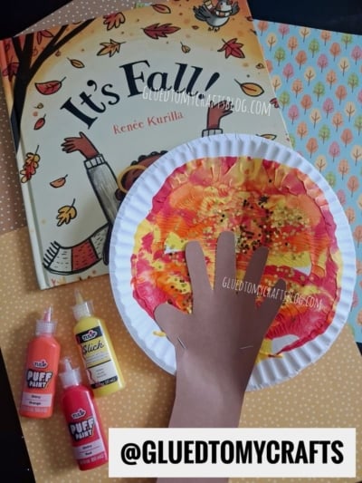 Children’s fall craft: a paper plate with painted autumn colors and a hand cutout as a tree, resembling a playful paint splat tree, next to bottles of puff paint and the book “It’s Fall!” by Renée Kurilla. Background includes dotted and tree-patterned paper.