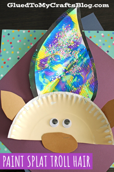 A troll craft made from a paper plate for the face, with paint splat troll hair using glittery leaf-shaped paper, googly eyes, and brown paper ears and nose, on a colorful background. Text reads: “PAINT SPLAT TROLL HAIR.”.