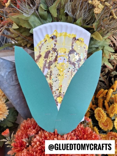A craft butterfly made from a painted coffee filter with green paper wings is displayed among orange and yellow artificial flowers, alongside a paint splat corn creation. The Instagram handle @GLUEDTOMYCRAFTS is visible at the bottom.