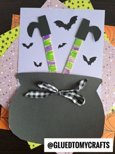 A paper craft displays a black cauldron with a bow, popsicle stick witch legs with stripes sticking out, and black bats flying above, all set on layered Halloween-themed papers.