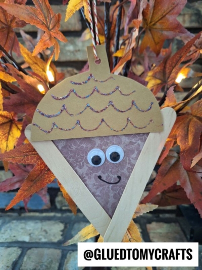 A handmade craft stick acorn ornament with googly eyes, made from paper and popsicle sticks, hangs on a string in front of fall leaves. @GLUEDTOMYCRAFTS appears at the bottom.