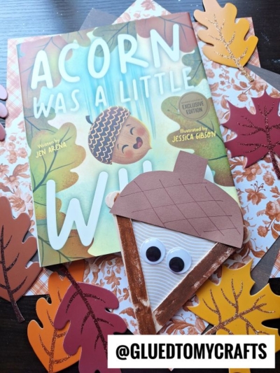 A children’s book titled “Acorn Was a Little Wild” is surrounded by colorful paper leaves. In the foreground, a handmade craft stick acorn with googly eyes adds a playful touch. The @GLUEDTOMYCRAFTS handle appears at the bottom.