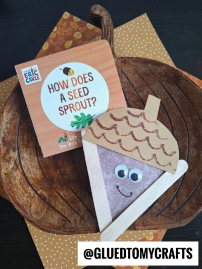 A children’s book titled How Does a Seed Sprout? sits on a wooden tray with a craft stick acorn made of paper, popsicle sticks, googly eyes, and glitter. The handle @GLUEDTOMYCRAFTS is at the bottom.