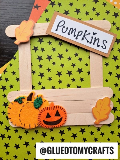 A popsicle stick pumpkin patch stand frame decorated with felt pumpkins, glitter, and leaf shapes rests on star-patterned paper. A sign labeled “Pumpkins” is attached to the frame, with @GLUEDTOMYCRAFTS written at the bottom.
