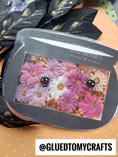 A fishbowl-shaped craft, reminiscent of a stained glass witch cauldron, filled with pink and white flowers, gold glitter, and two small black pom-pom creatures with googly eyes. The background has leaves and polka dot paper. Text reads @GLUEDTOMYCRAFTS.