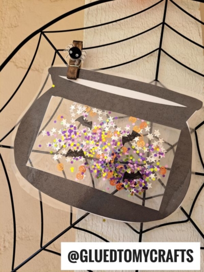 A paper fishbowl with confetti, bat shapes, and star stickers is clipped to a spiderweb decoration, creating a whimsical scene reminiscent of a stained glass witch cauldron bubbling with magic. A small spider clip is at the top. @GLUEDTOMYCRAFTS appears at the bottom.