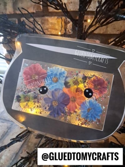 A colorful paper fishbowl craft with pressed flowers and glitter, decorated with two black cartoon fish, hangs on a black branch. Reminiscent of a stained glass witch cauldron in its vibrant hues and magical charm, the handle @GLUEDTOMYCRAFTS is displayed in bold at the bottom.