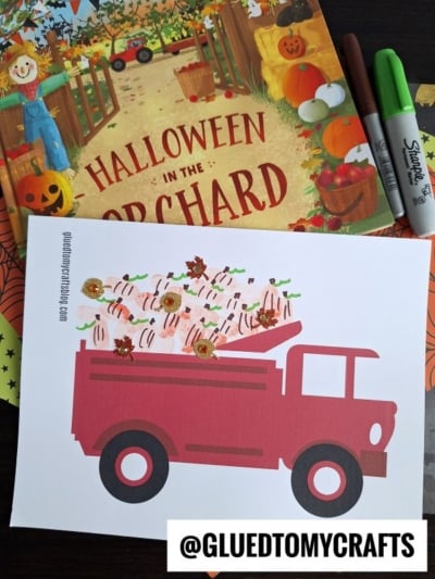 A Halloween-themed craft scene with a book titled Halloween in the Orchard, Sharpie markers, and a paper craft of a red truck carrying thumbprint pumpkins. The @GLUEDTOMYCRAFTS username is visible at the bottom.