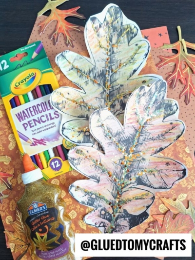 Assorted fall-themed craft supplies, including watercolor pencils, gold glitter glue, autumn leaves, a watercolor leaf, and two large paper leaves decorated with colorful paint and glitter, arranged on a patterned background.
