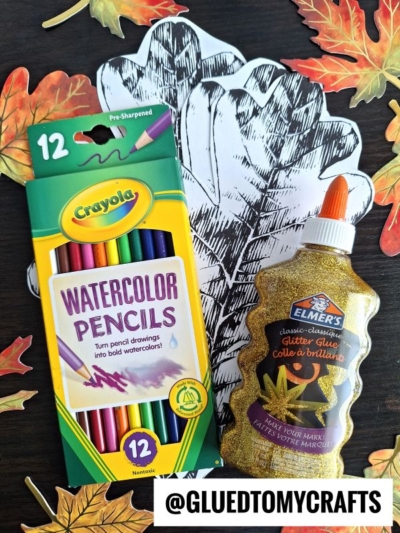 Watercolor pencils, a bottle of gold glitter glue, and black-and-white paper leaf cutouts are surrounded by artificial autumn leaves and vibrant watercolor fall leaves on a dark tabletop. @GLUEDTOMYCRAFTS text is shown at the bottom.